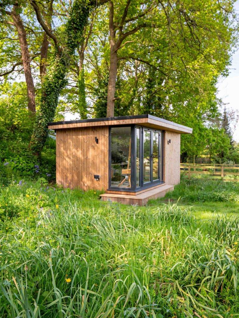 Small wooden cabin with large glass doors situated among tall grass and surrounded by green trees. For a free visit, contact us today!